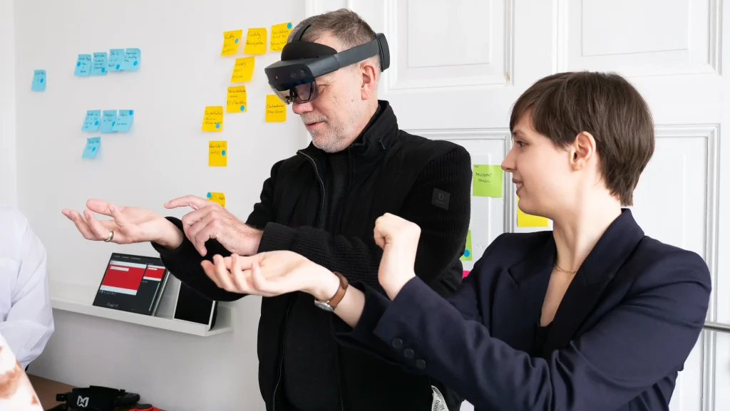 Eine Person trägt eine Mixed-Reality-Brille, während eine weitere Person die Gestensteuerung zu der Brille erklärt