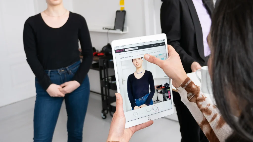 Eine Person richtet ein Tablet mit Kamera auf eine andere Person. Dieser wird Schmuck in Augmented Reality angelegt.