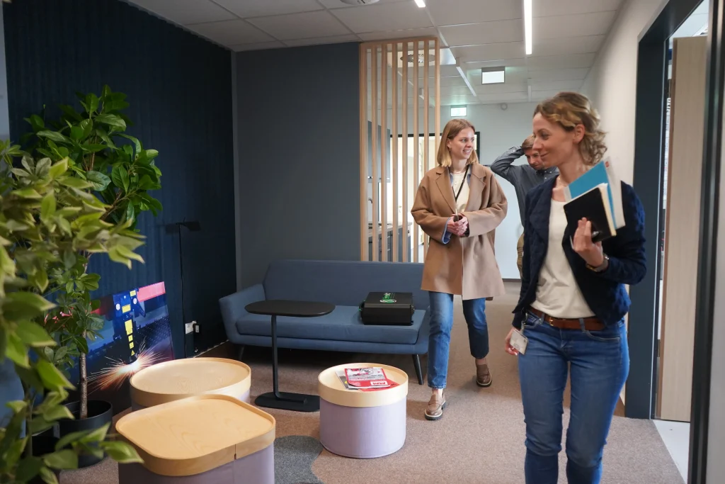 Drei Personen laufen durch ein Bürogebäude. Zu sehen ist eine gemütliche Sitzecke mit Sofa und Arbeitsfläche.