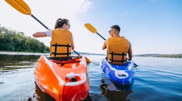 Ein Pärchen, die zusammen auf einem Fluss Kayak fahren. Eine Person ist weibliche und fährt ein orangenes Kayak. Die andere Person ist männlich und fährt ein blaues Kayak. Beide tragen eine orangene Schwimmweste.