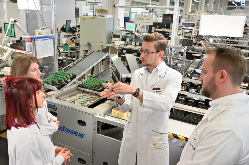 Das Bild zeigt vier Personen in einem Fertigungsumfeld. Zwei Frauen und zwei Männer stehen um eine Arbeitsstation herum und analysieren das Produkt der Fertigungslinie.