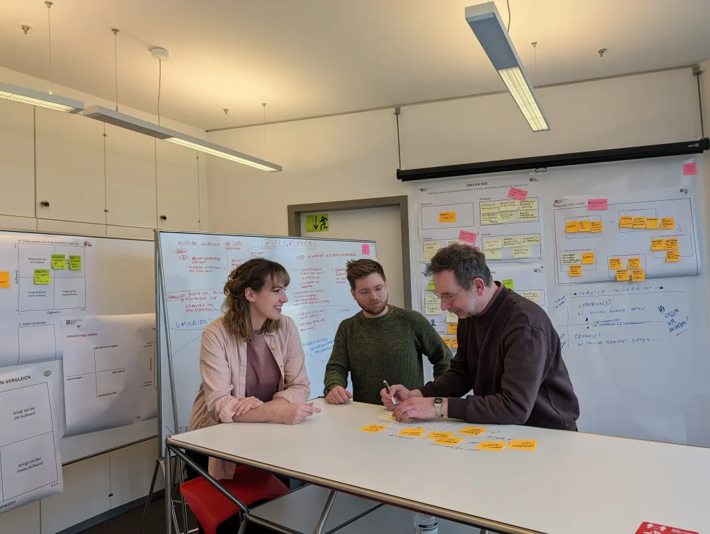 Drei Personen stehen in einem Workshopraum an einem Stehtisch und besprechen die Post-its, die vor ihnen auf dem Tisch liegen.