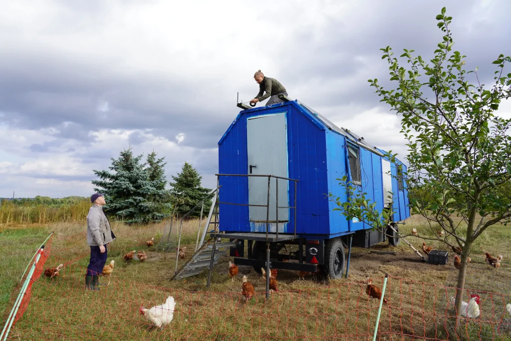 Ein mobiler Hühnerwagen im Hintergrund. Auf dem Wagen sitzt ein Entwickler und installiert Technik auf dem Dach. Der Bauer steht neben dem Wagen und schaut zu. Hühner sind im Vordergrund auf der Wiese zu sehen.