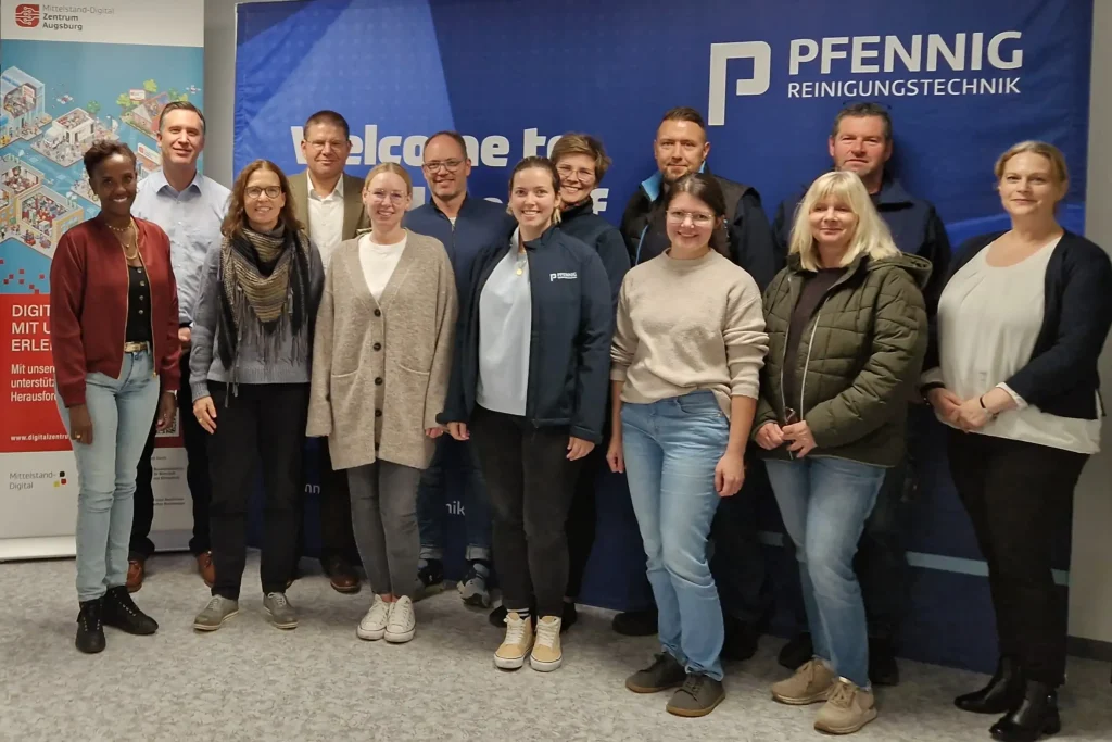 Gruppenfoto der Workshop-Teilnehmer:innen der PFENNIG Reinigungstechnik GmbH und von ibi research vor dem Roll-up des Mittelstand-Digital Zentrums Augsburg. Innenraum.