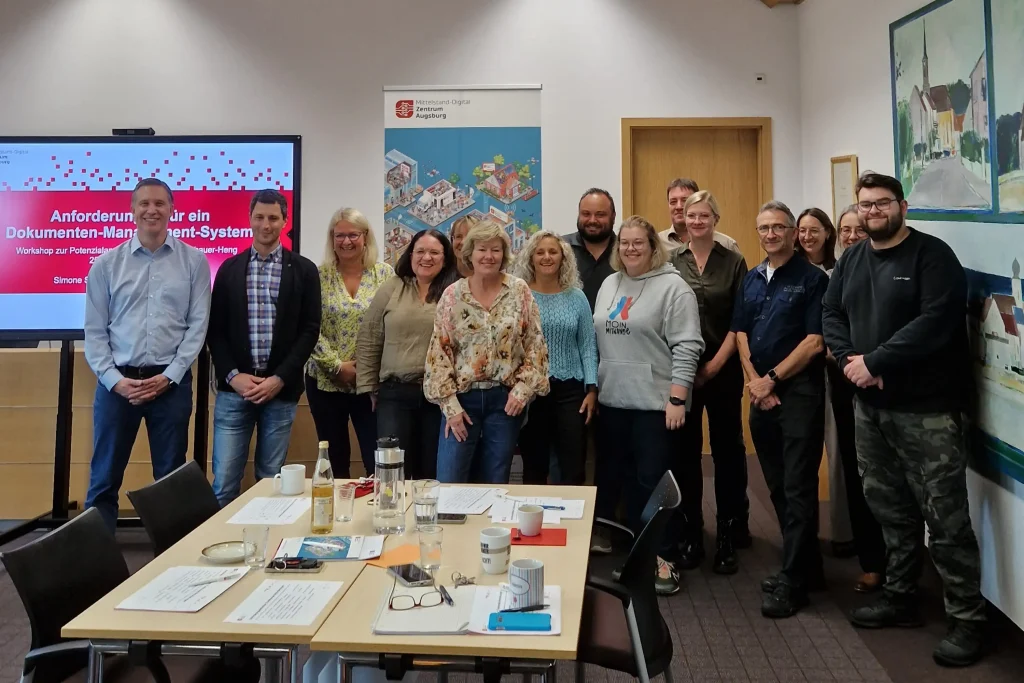 Teilnehmer:innen des Workshops zur Anforderungserhebung. Gruppenfoto der Mitarbeitenden des Markts Postbauer-Heng und von ibi research vor dem Roll-up des Mittelstand-Digital Zentrums Augsburg. Innenraum.