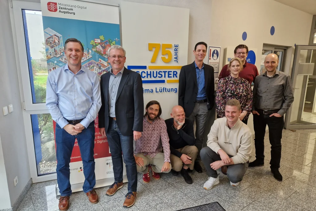 Gruppenfoto der Workshop-Teilnehmer:innen der Schuster Klima Lüftung GmbH & Co. KG und von ibi research vor dem Roll-up des Mittelstand-Digital Zentrums Augsburg. Innenraum.