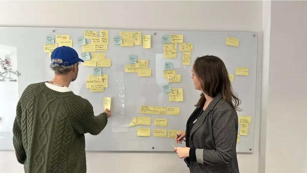 Zwei Menschen stehen vor einem Whiteboard mit Haftnotizen und clustern diese zu übergreifenden Themen