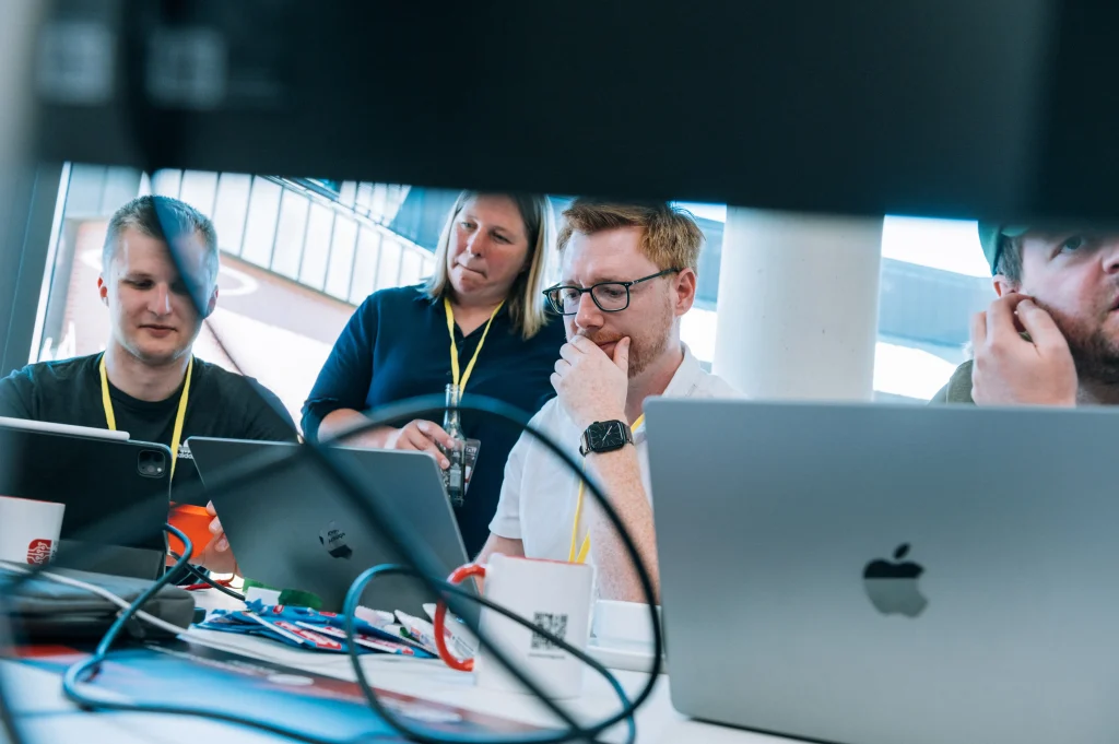 Mehrere Personen arbeiten gemeinsam an einem Tisch mit Laptops, Kabeln und konzentrierter Miene