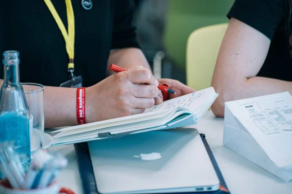 Eine Person schreibt mit einem roten Stift in ein geöffnetes Notizbuch. Auf dem Tisch liegen ein MacBook, eine Wasserflasche, ein Papierbeutel und Stifte. Neben der schreibenden Person sitzt eine weitere Person.