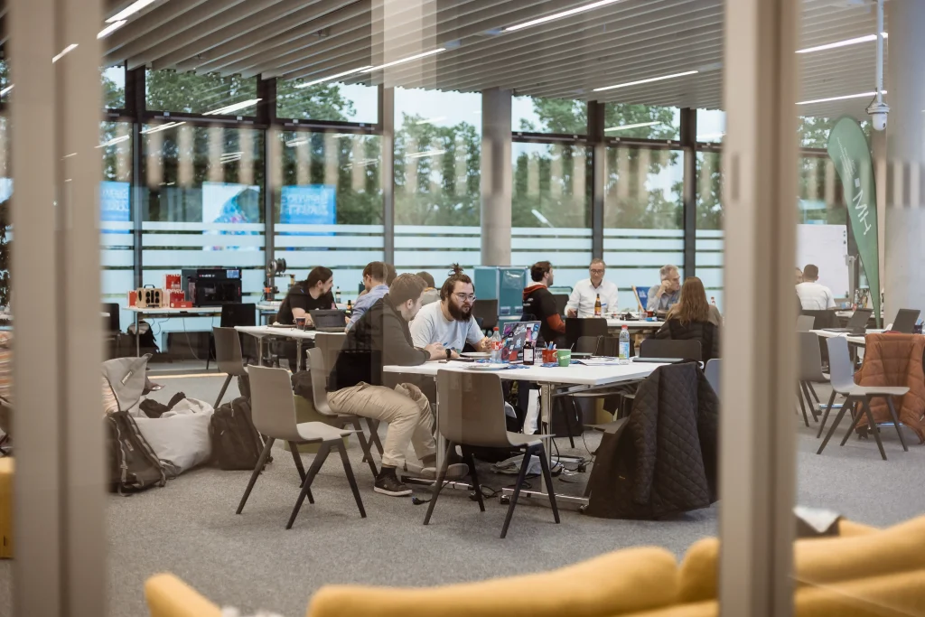 Das Bild zeigt einen modernen, hellen Arbeitsraum mit großen Fenstern, in dem mehrere Menschen an Tischen konzentriert mit Laptops und technischen Geräten arbeiten im Rahmen eines Team-Workshops.