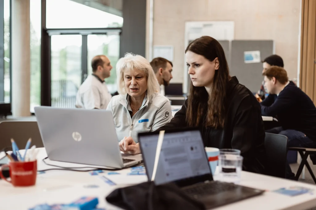Das Bild zeigt zwei Frauen an einem Tisch, die konzentriert auf einen Laptop schauen und gemeinsam arbeiten, während im Hintergrund weitere Personen in Gespräche oder Computerarbeit vertieft sind – die Umgebung wirkt modern und kooperativ