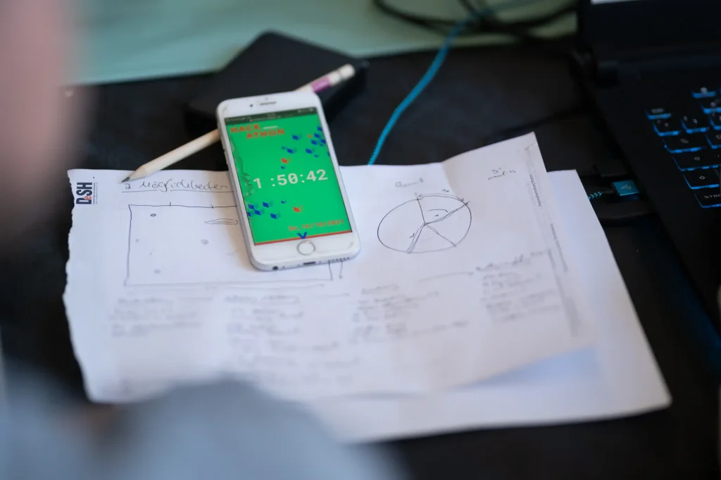 Das Bild zeigt ein Smartphone, das auf einem Blatt mit handschriftlichen Notizen und Skizzen liegt. daneben befinden sich ein Stift und ein Laptop – alles deutet auf eine konzentrierte Arbeits- oder Projektsituation hin.