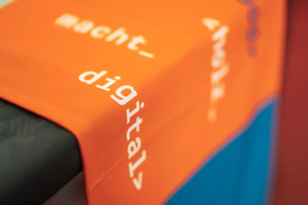 Ein orange farbenes Banner, was über eine Tischkante hängt. In weißer Druckschrift ist deutlich das Wort