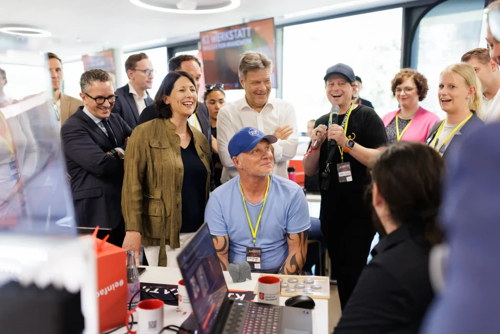 Eine Gruppe von Menschen steht lachend und interessiert um einen Tisch mit Computern und Technik, während eine Person im blauen T-Shirt und blauer Kappe im Mittelpunkt sitzt.