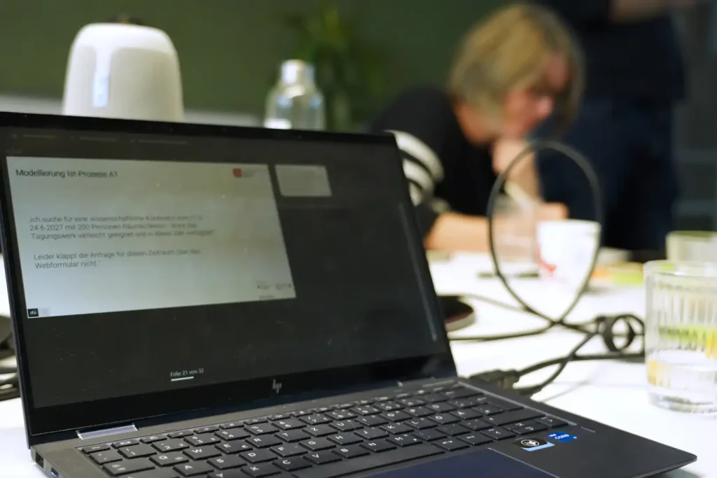 Auf dem Laptop ist eine Beispielanfrage für eine Konferenz zu sehen, anhand derer der bestehende Bearbeitungsprozess analysiert wird.