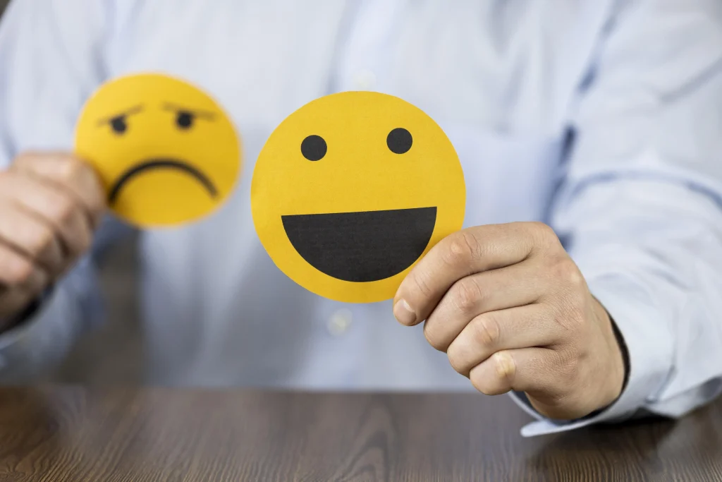 Eine Person, die einen fröhlichen und traurigen Smiley in die Kamera hält. Der fröhliche ist im Vordergrund.