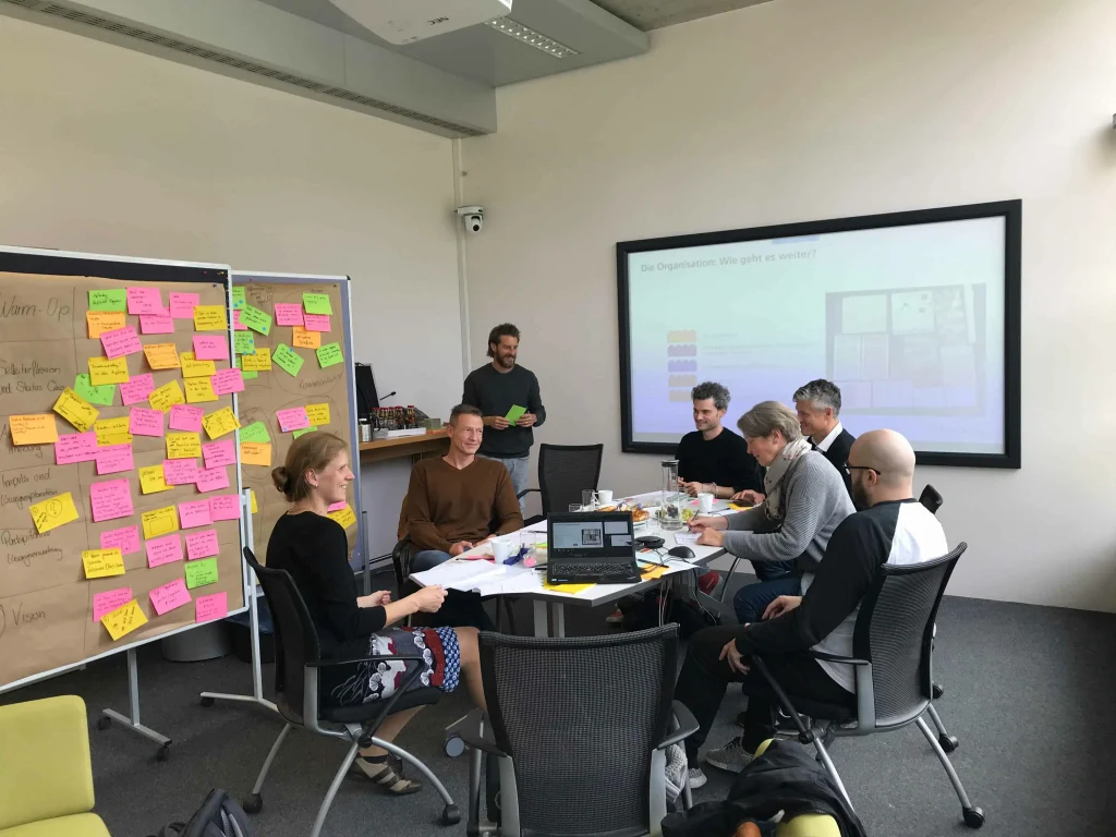 Workshopsituation, beteiligte Personen sitzen am Tisch und diskutieren. Die moderierende Person steht im Hintergrund zwischen Stellwänden mit Haftnotizen und dem Bildschirm, wo die aktuelle Folie sichtbar ist.