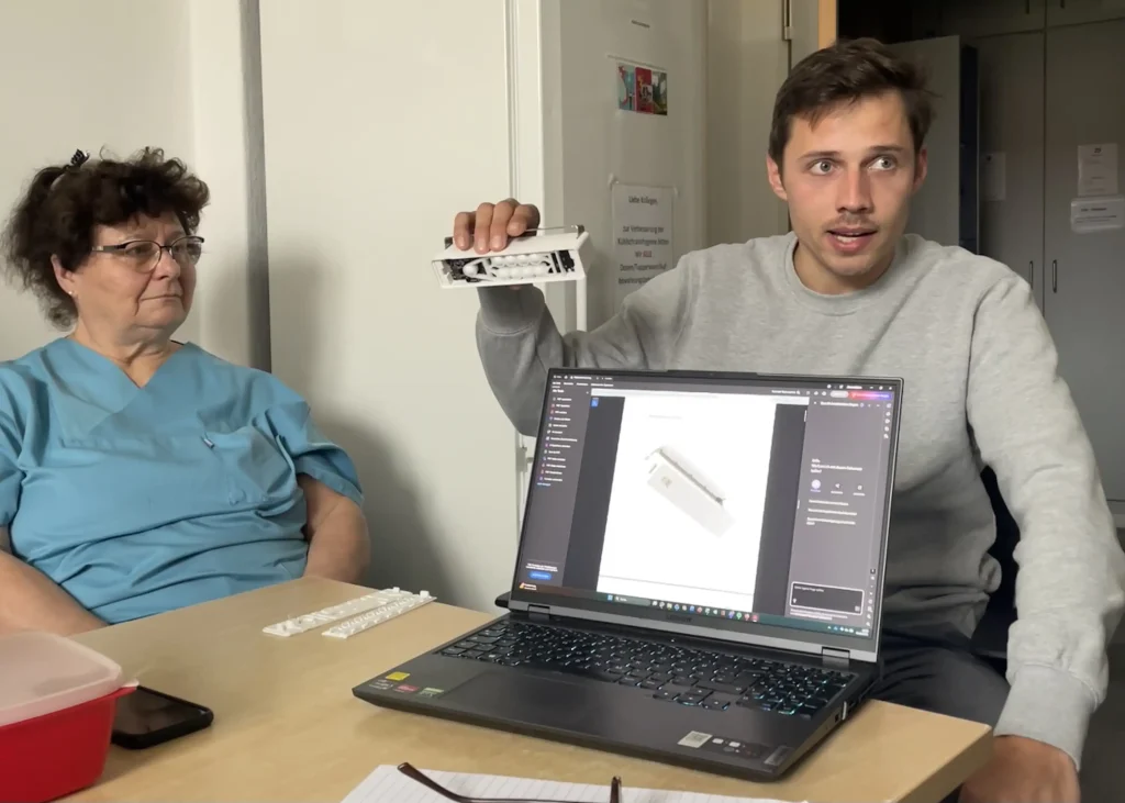 Zwei Personen sitzen an einem Besprechungstisch. Eine Person präsentiert ein weißes technisches Bauteil und hält es in die Kamera, während auf einem geöffneten Laptop eine digitale Darstellung desselben Objekts zu sehen ist. Die zweite Person beobachtet d