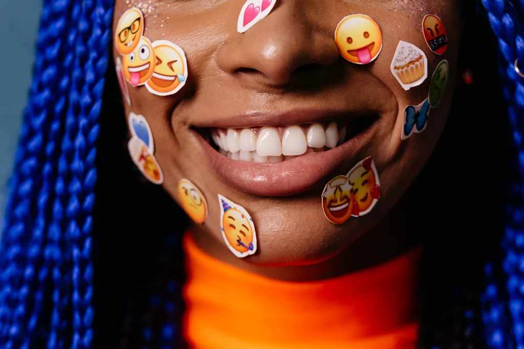 Nahaufnahme einer lächelnden Person mit blauen Zöpfen und bunten Emoji-Aufklebern im Gesicht, die verschiedene Emotionen darstellen. Sie trägt ein leuchtend oranges Oberteil.