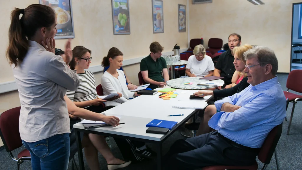 In einem Abschlussworkshop sitzen mehrere Teilnehmende an einem großen Tisch und tauschen sich aus. Eine Frau steht vorn und moderiert die Diskussion, während die übrigen Personen Notizen machen und mit bunten Haftnotizen arbeiten.