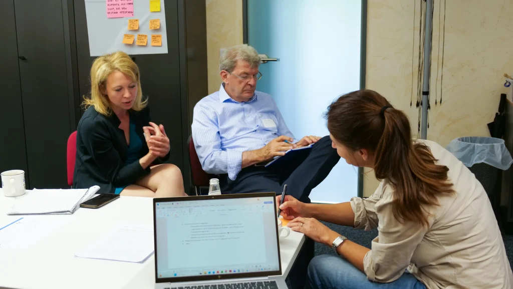 Drei Personen arbeiten in einer kleinen Workshopgruppe an einem Tisch. Eine Frau erläutert einen Punkt mit Gesten, während ein Mann Notizen auf einem Klemmbrett macht. Im Vordergrund schreibt eine weitere Frau neben einem geöffneten Laptop. Auf dem Tisch