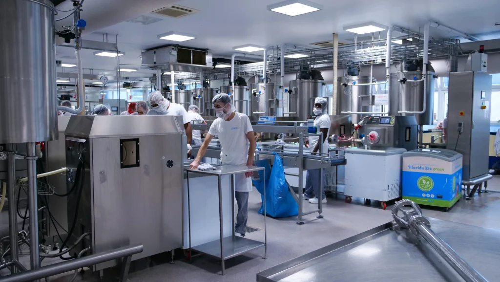 Mehrere Mitarbeitende in weißer Arbeitskleidung, Haarnetzen und Gesichtsmasken arbeiten in einer hell beleuchteten Produktionshalle. Zwischen großen Edelstahlbehältern, Rohrleitungen und Maschinen bedienen sie Anlagen zur Herstellung und Verarbeitung von
